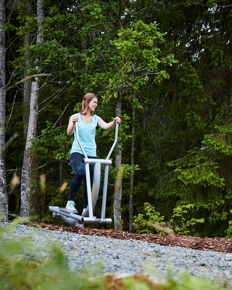 A woman exercises on an outdoor elliptical exercise machine, she is surrounded by nature and trees.